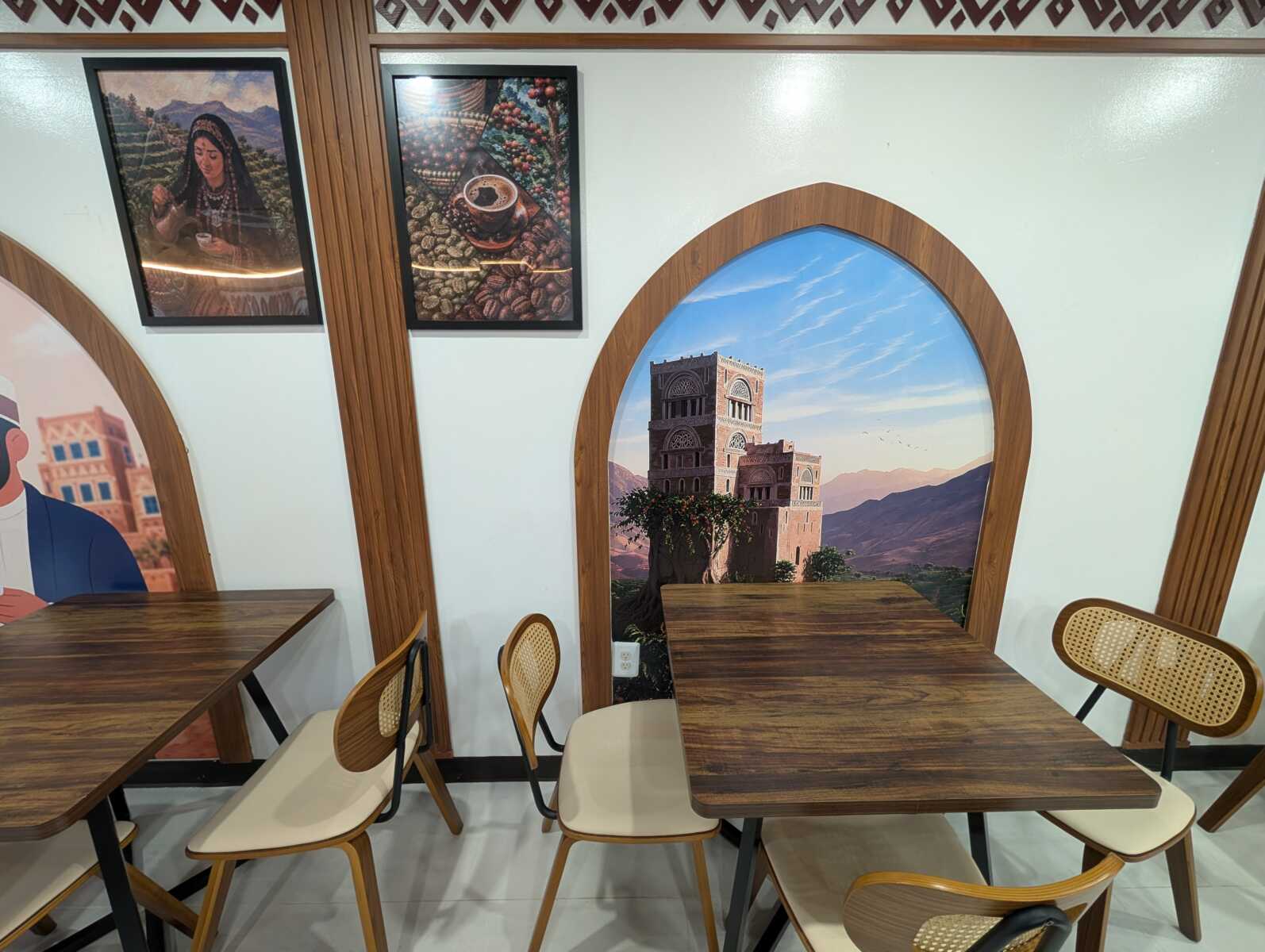 Cafe seating area with dark wood tables and cream chairs, framed wall art, and arched window mural depicting a historic tower against a desert landscape.