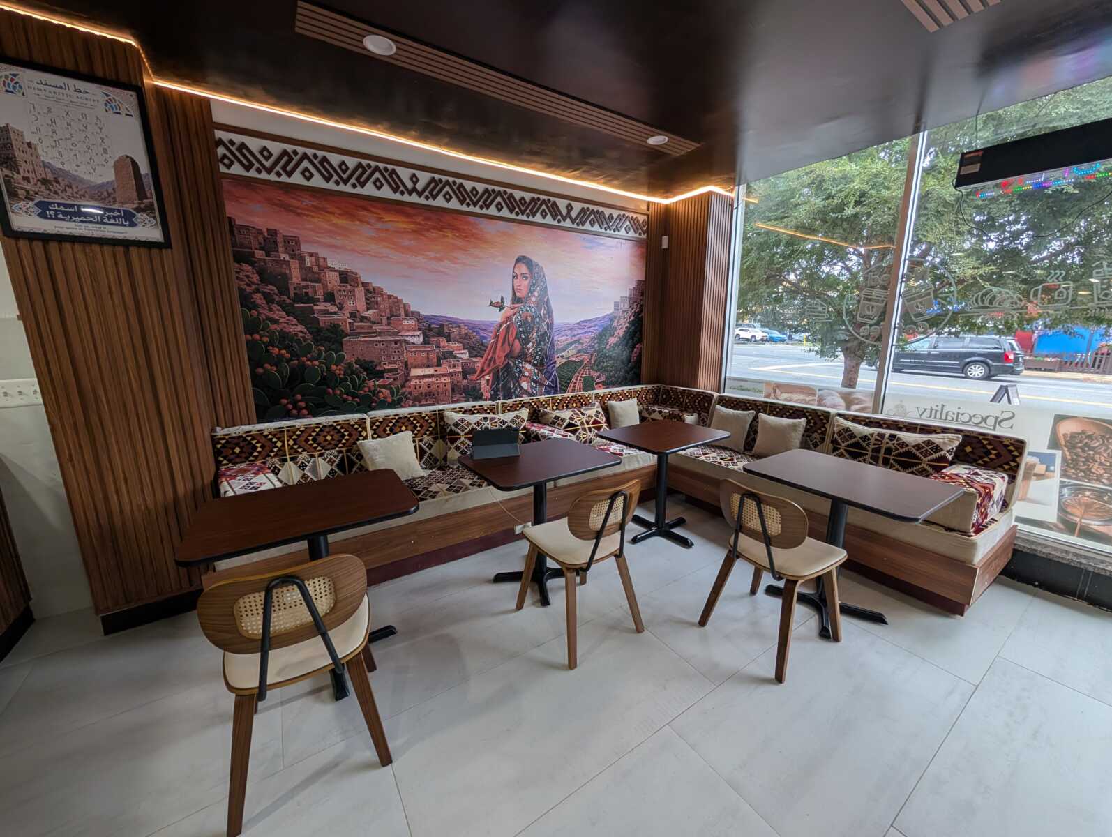 Cozy cafe interior with a large mural of a woman in traditional attire, surrounded by patterned brown seating and wooden accents. A long bench and small tables line the wall by a window.