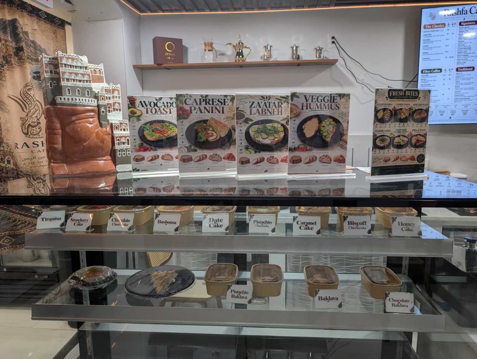 Cafe display case with posters advertising Avocado Toast, Caprese Panini, Za'atar Labneh, and Veggie Hummus, plus cakes behind glass.