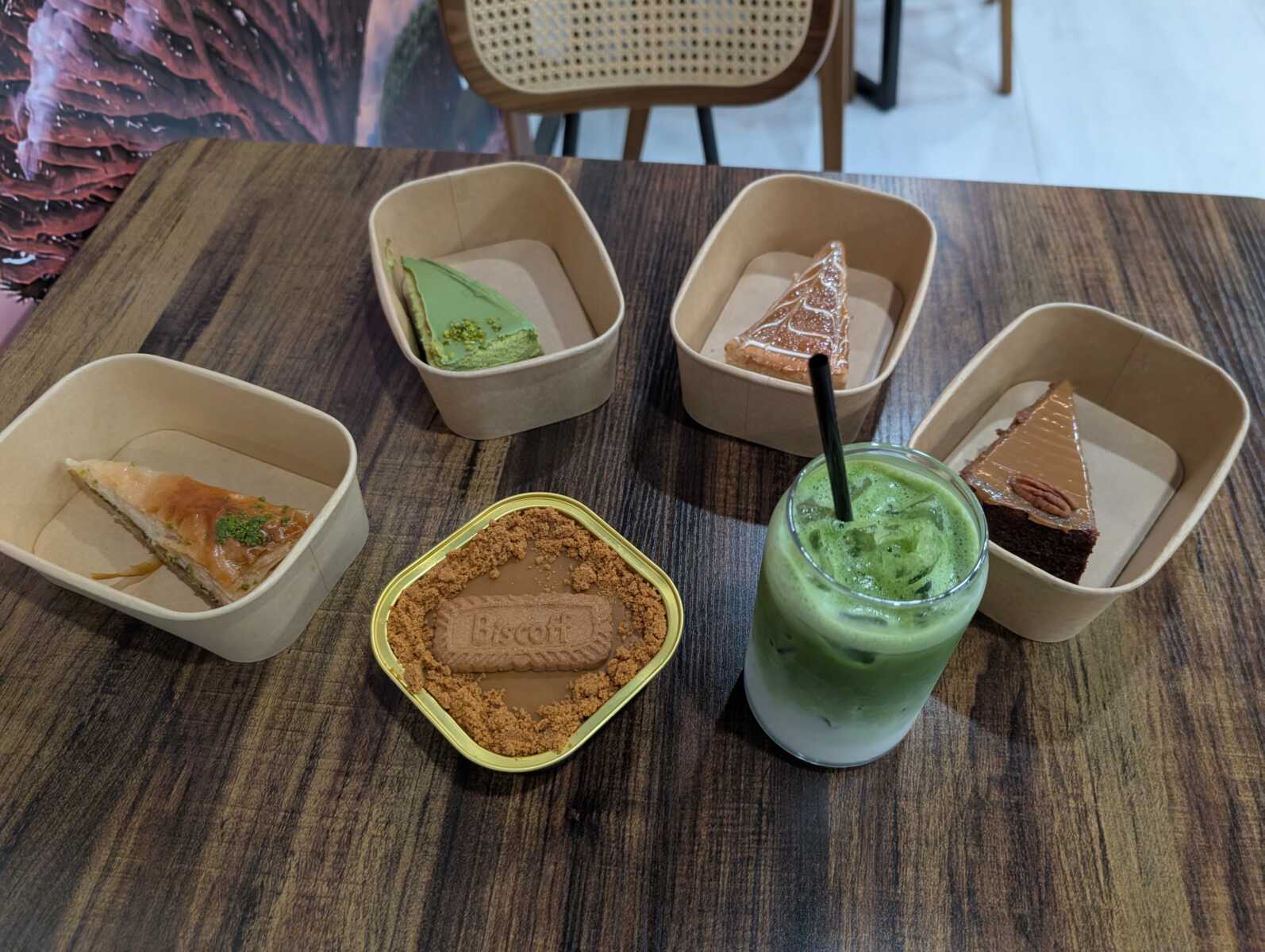Assorted mini cakes on a wooden table: green matcha slice, two chocolate-topped slices, and a crumb-tinished tart, plus a green iced drink with a black straw nearby.