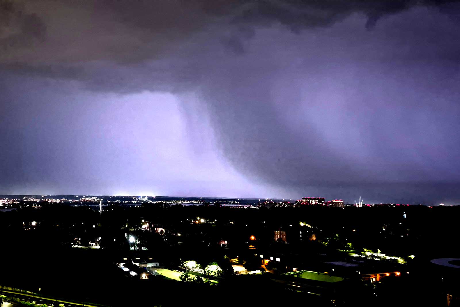Lightning and rain during Thursday night's storm (courtesy James Mahoney) | ARLnow Nighttime cityscape under a massive thunderstorm with a purple-tinged shelf cloud stretching across the horizon and bright city lights below.