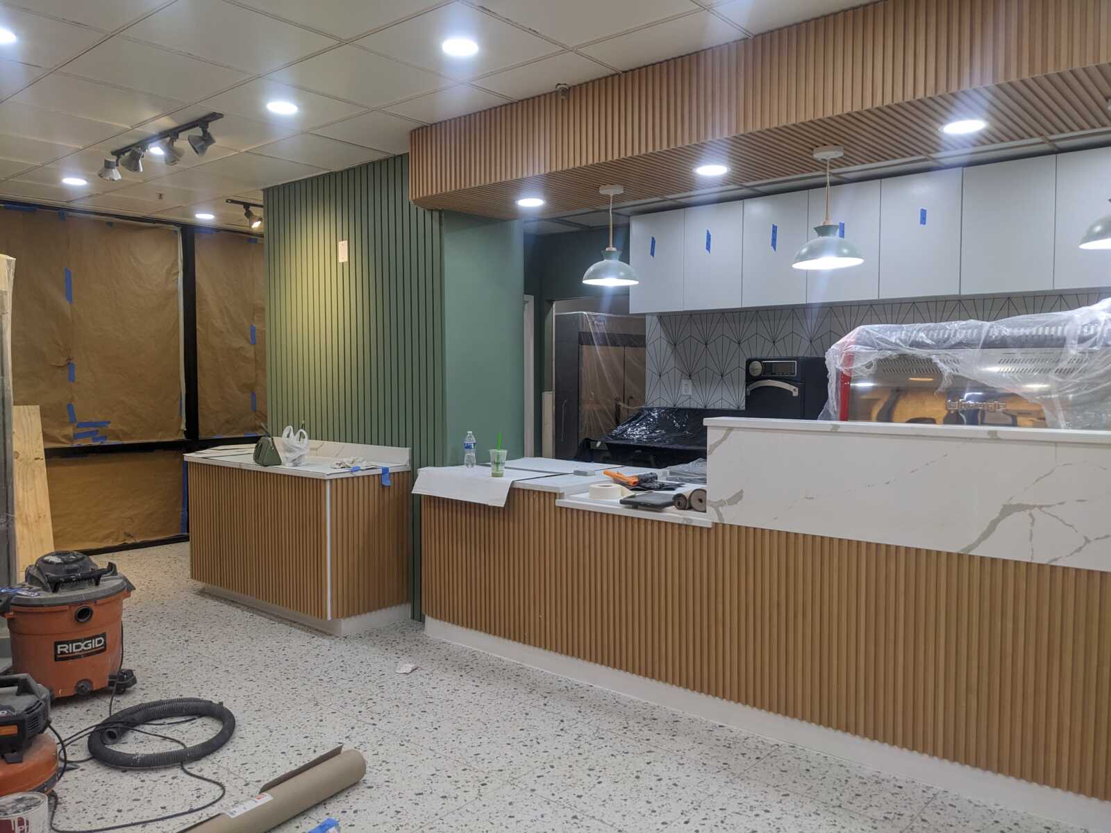 Cafe counter under renovation with wooden front panels, green wall, and plastic-covered equipment in background.