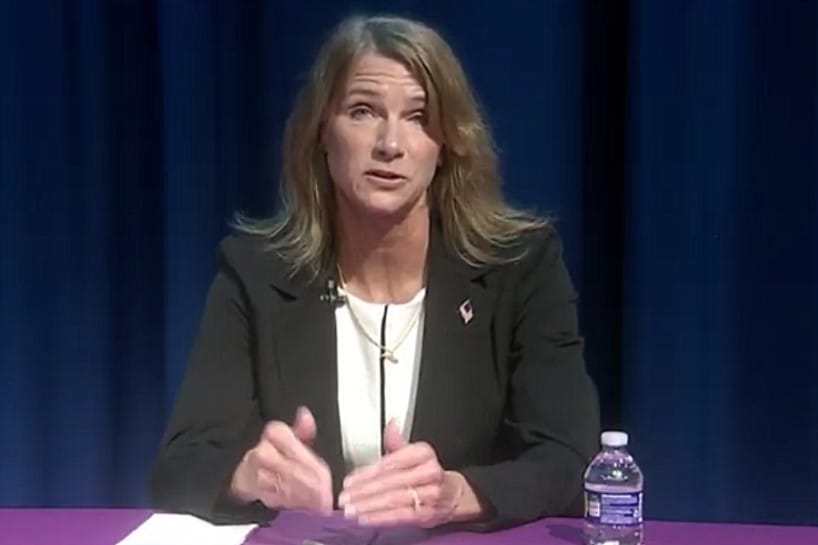 Woman in a black blazer speaks at a panel, gesturing with her hands, with a mic clipped to her lapel and a water bottle on a purple table in front of her.