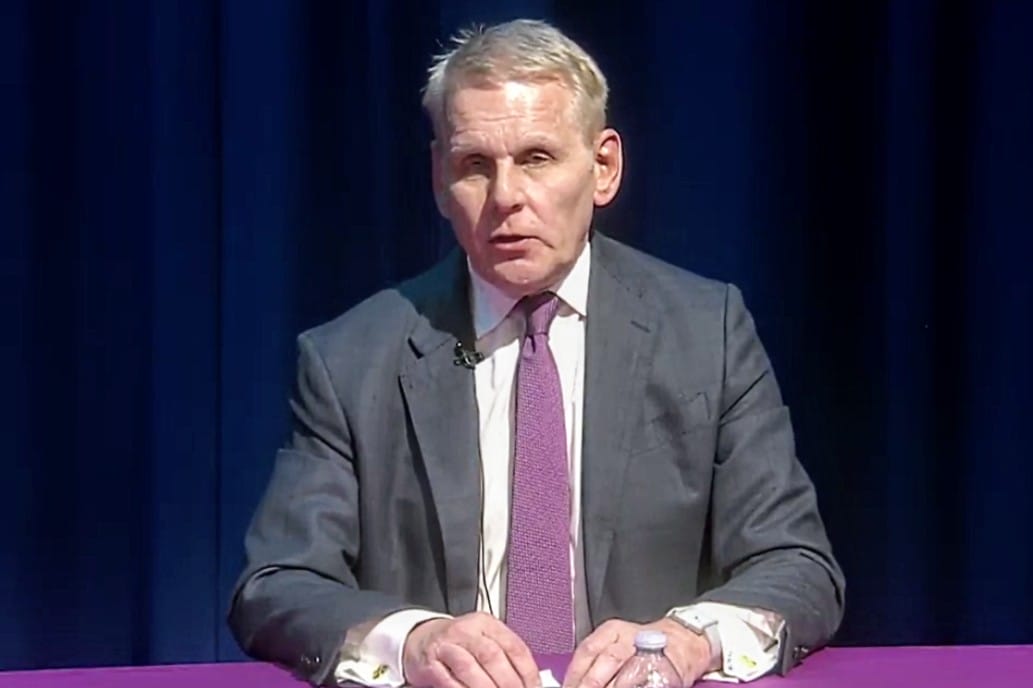 Man in a gray suit and pink tie sits at a pink table with a lapel microphone in front of a dark blue curtain backdrop.