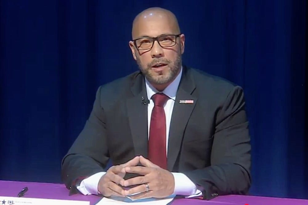Bartender? No. A man in a dark suit and burgundy tie sits at a conference table, speaking with hands folded, wearing glasses and a lapel pin against a blue backdrop.