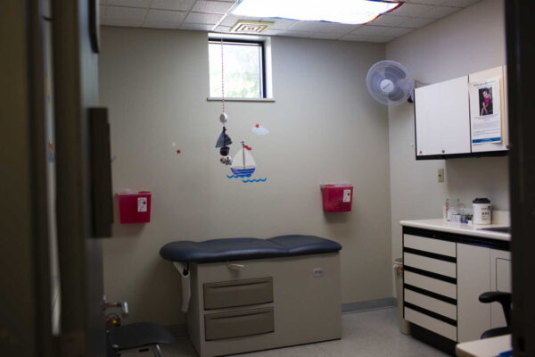 Medical exam room with a blue padded examination table, wall sailboat decals, and pink sharps containers; cabinets and medical supplies visible to the right.