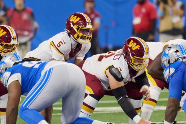 American football players from two teams line up at the line of scrimmage, wearing white maroon and blue uniforms, ready for a play.