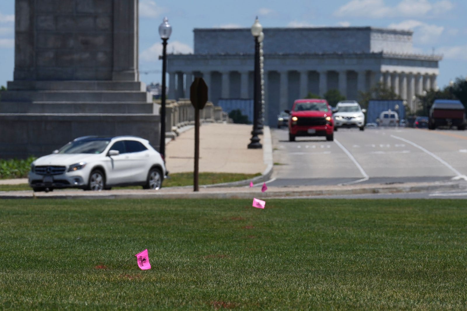 Wide street scene near a large neoclassical monument; pink flags dot the grassy median as cars drive by on a sunny day