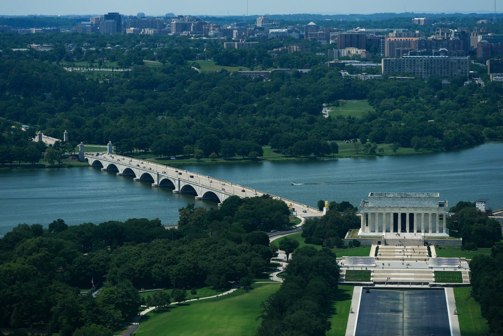 Aerial view of the Lincoln Memorial and a long arched bridge spanning a river, with a green park and city skyline in the distance.