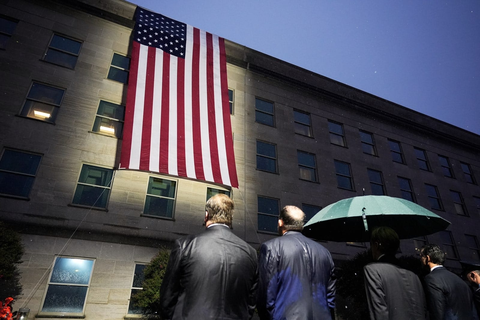 Large American flag hung vertically on a gray office building at dusk, with suited people in foreground and a green umbrella nearby.