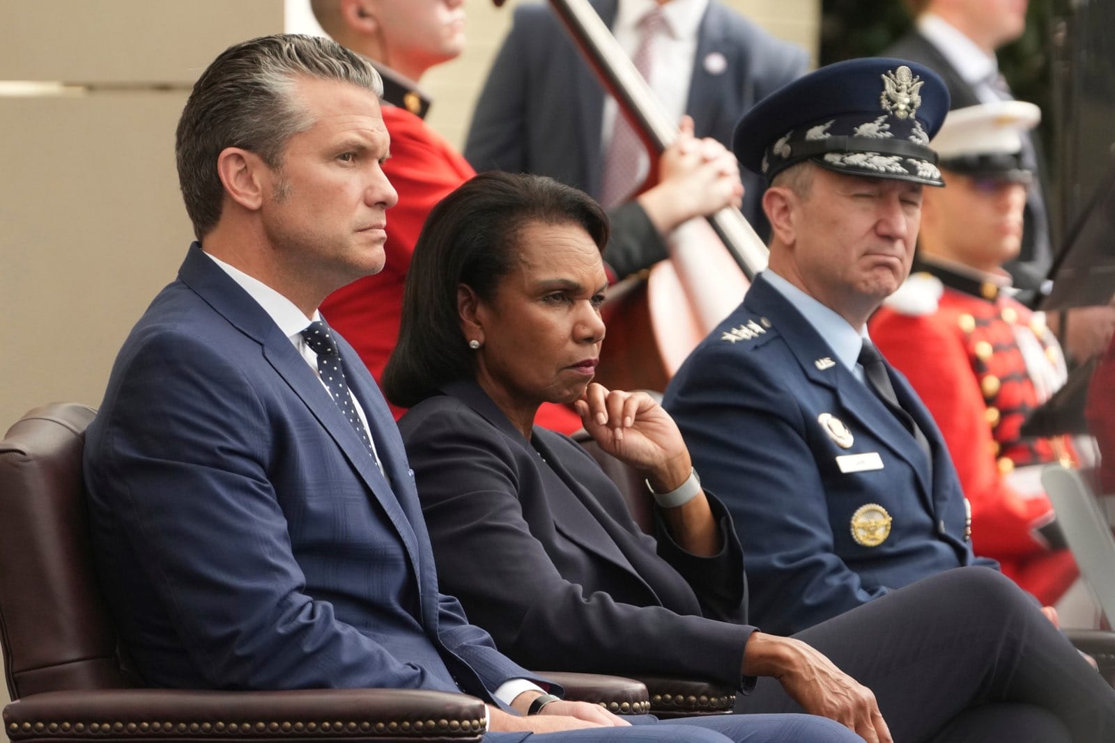 Three officials seated at a formal ceremony: a man in a blue suit, a woman in a dark blazer with her hand near her chin, and a uniformed officer in dress blues, with ceremonial guards in the background.