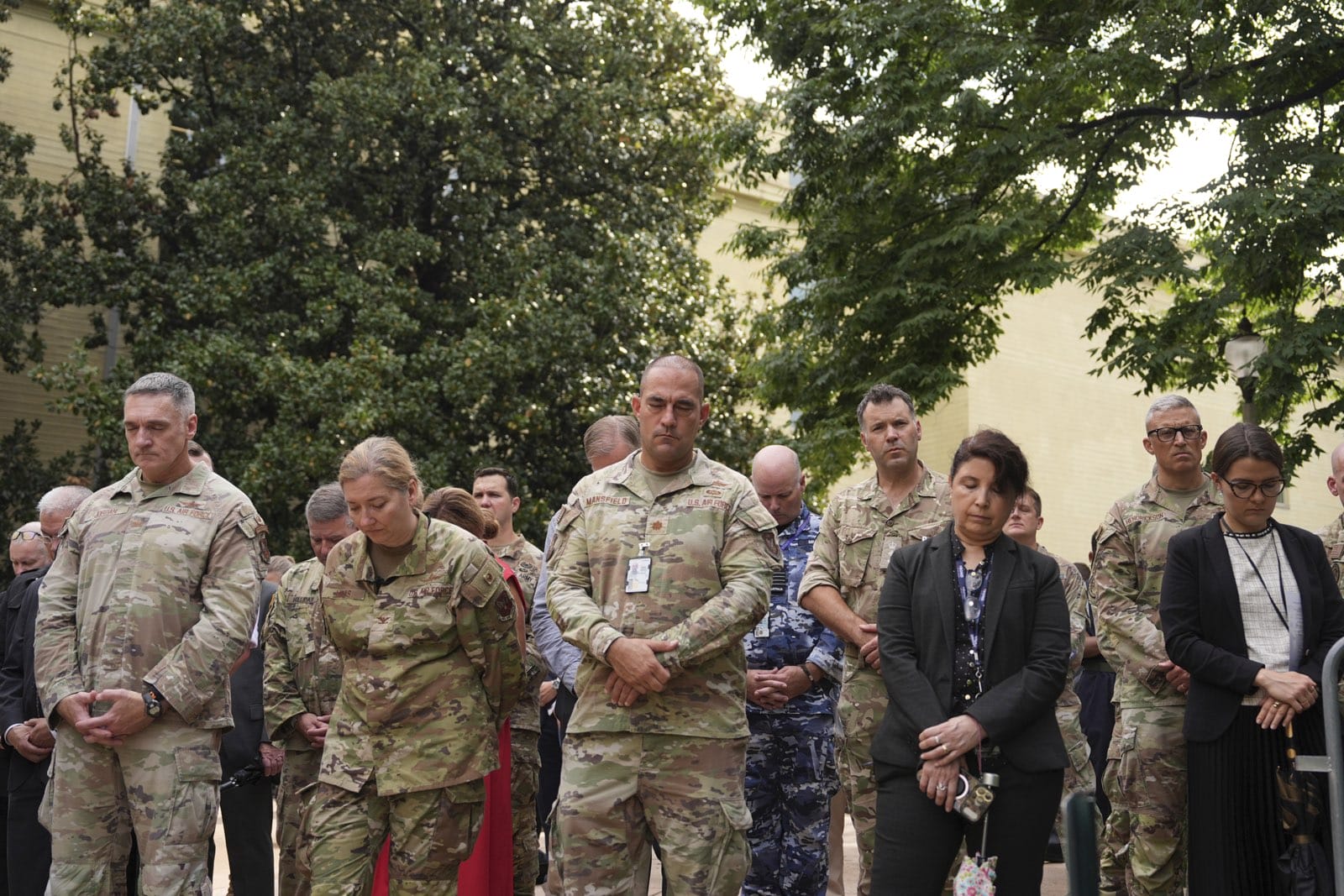 Group of soldiers and civilians standing with heads bowed outdoors during a moment of silence, wearing camouflage uniforms and formal attire.