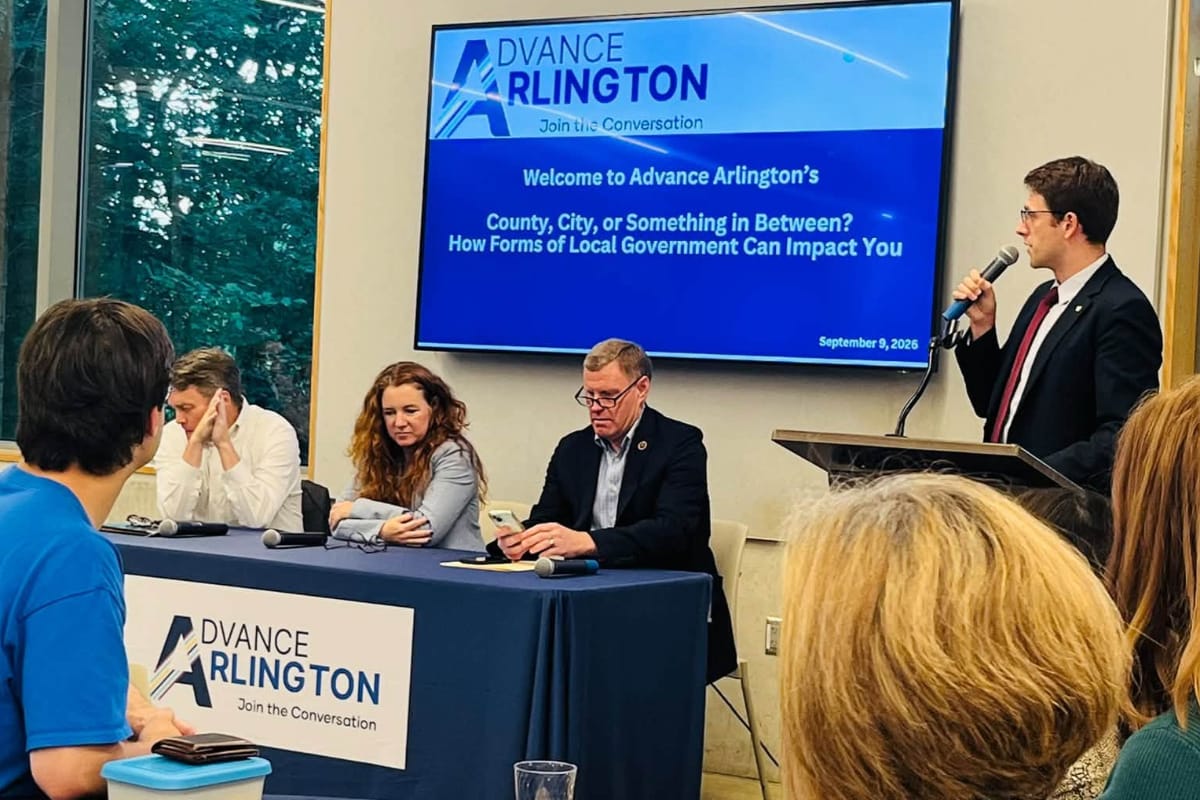 Advance Arlington program on governance structures across Northern Virginia (courtesy Toni Andrews) | ARLnow Panel discussion at Advance Arlington: four speakers seated at a blue-draped table, a man at a podium speaks into a microphone while a screen behind shows the event title.