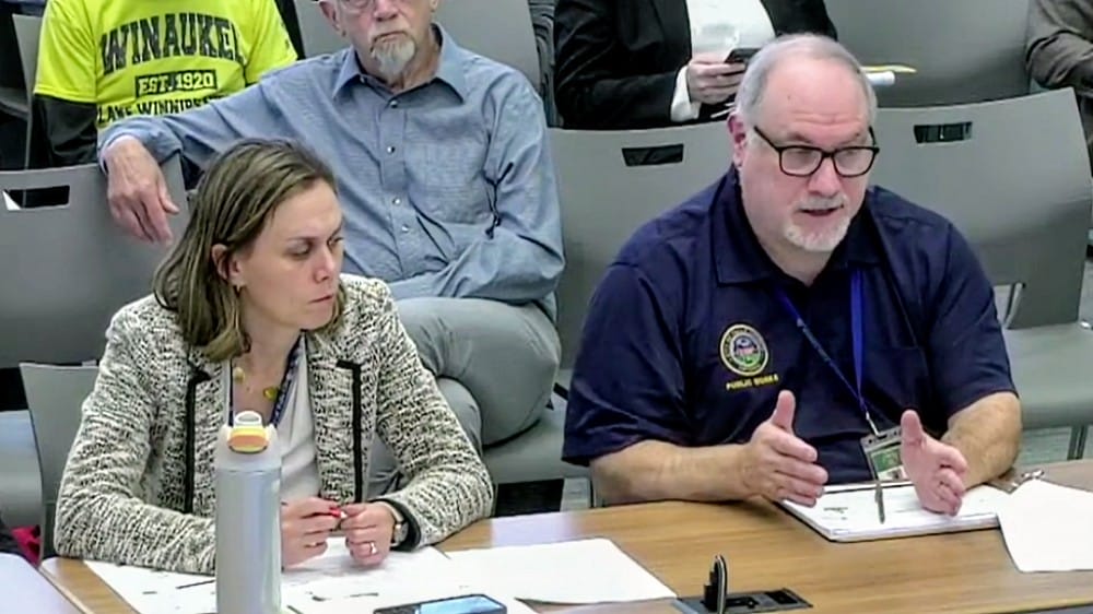 Amanda Stout Brain and Scott Freda discuss multimodal improvements on Broad Street (screenshot via City of Falls Church | ARLnow Two adults sit at a table in a meeting; the woman on the left looks down, the man on the right speaks with a notebook in front of him and a lanyard nearby.
