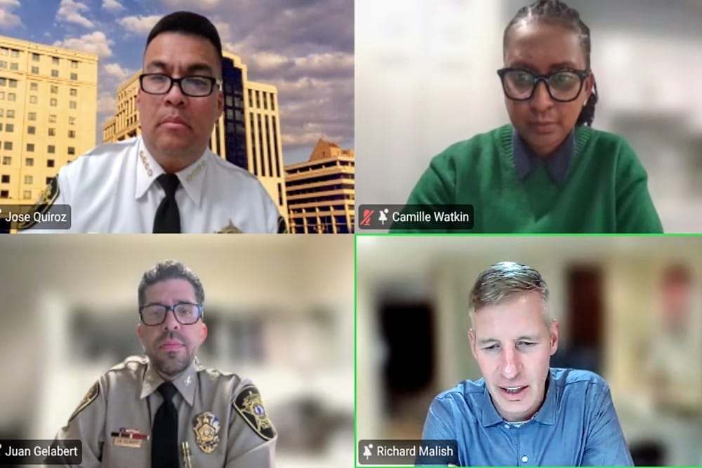 Four-person video conference grid: top-left man in white shirt and tie with city buildings behind, top-right woman in green sweater, bottom-left man in uniform, bottom-right man in blue shirt indoors.