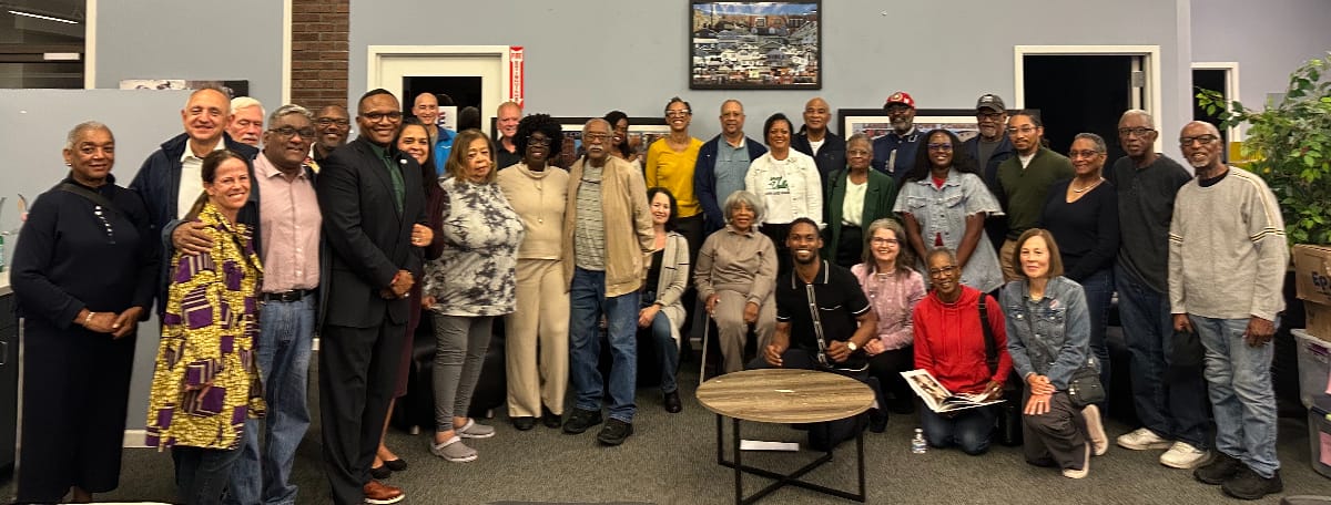 Attendees at arts fund-raiser (courtesy Wilma Jones) | ARLnow Large diverse group of adults posing for a group photo in an office setting, some standing and others kneeling around a round table.