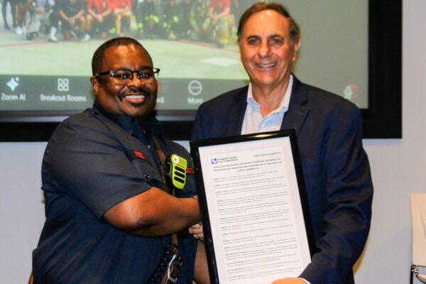 Two men shake hands while presenting a framed certificate at an awards event; one in a dark uniform smiles warmly.