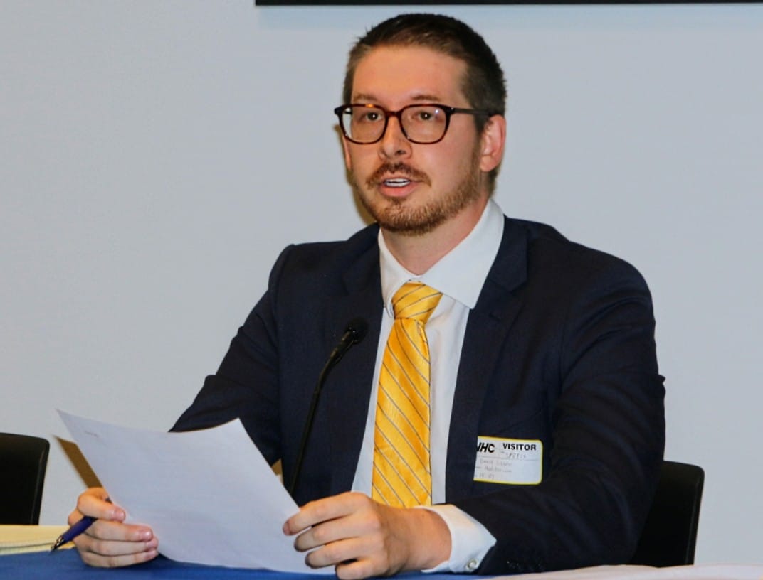 Man in a navy suit and yellow striped tie sits at a table, reading a document with a microphone in front.