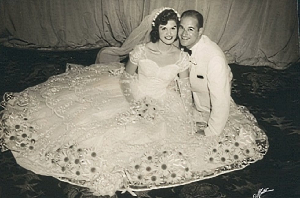Cynthia and Mort Zetlin wedding photo (courtesy Arlington Optimist Club) | ARLnow Bride in a voluminous, flower-trimmed wedding gown sits beside a groom in a white suit, posing on a dance floor.