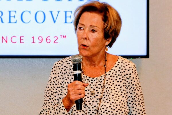 Woman speaking into a handheld microphone at a conference, wearing a white dotted blouse and gold necklace, with a screen in the background showing partial text.