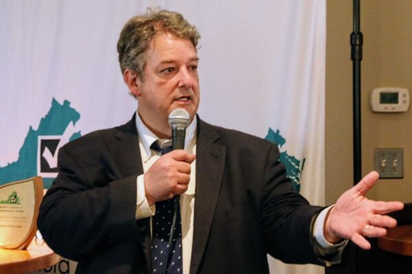 Man in a dark suit speaking into a handheld microphone at a panel or event, gesturing with his right hand