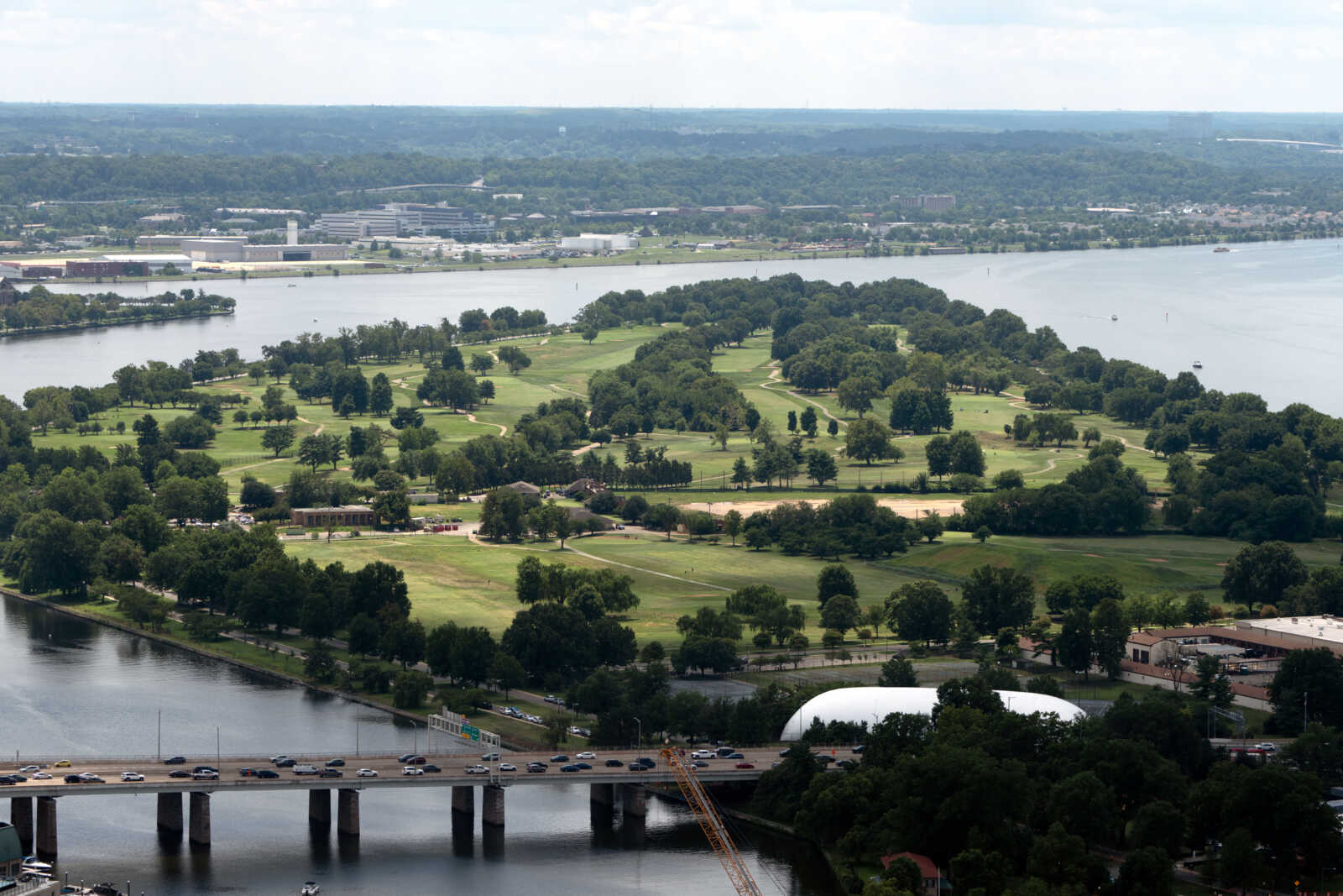 Aerial view of a tree-lined golf course on a narrow island in a wide river, with a bridge and highway below.
