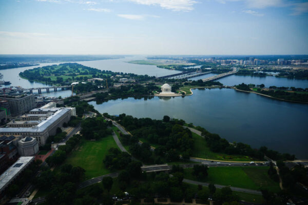 Aerial view of the Jefferson Memorial on the Tidal Basin, surrounded by water, greenery, and distant city bridges and skyline.