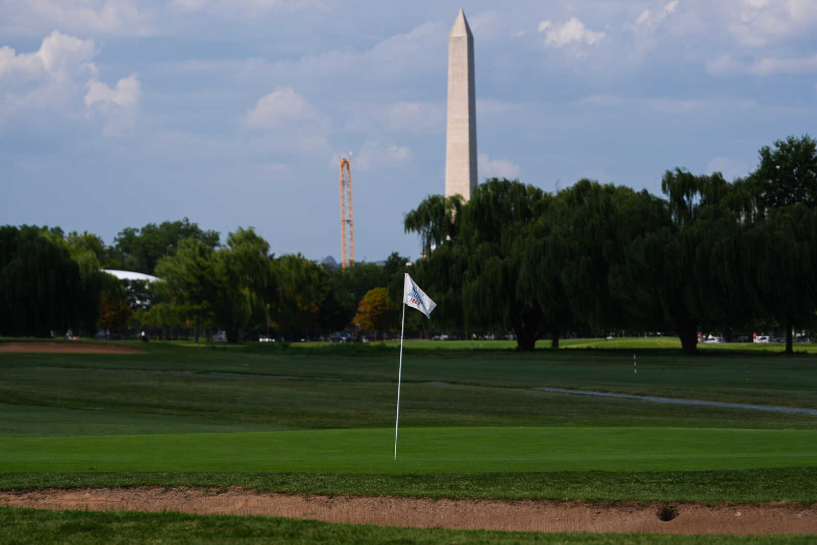 Golf green with a flagstick and hole, sand bunker in the foreground, and the Washington Monument visible beyond the trees.