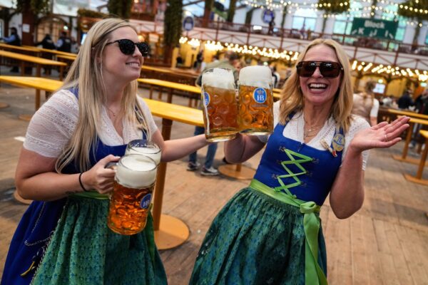 Two women in dirndls raise frothy beer mugs in a toast at an outdoor beer hall under string lights.