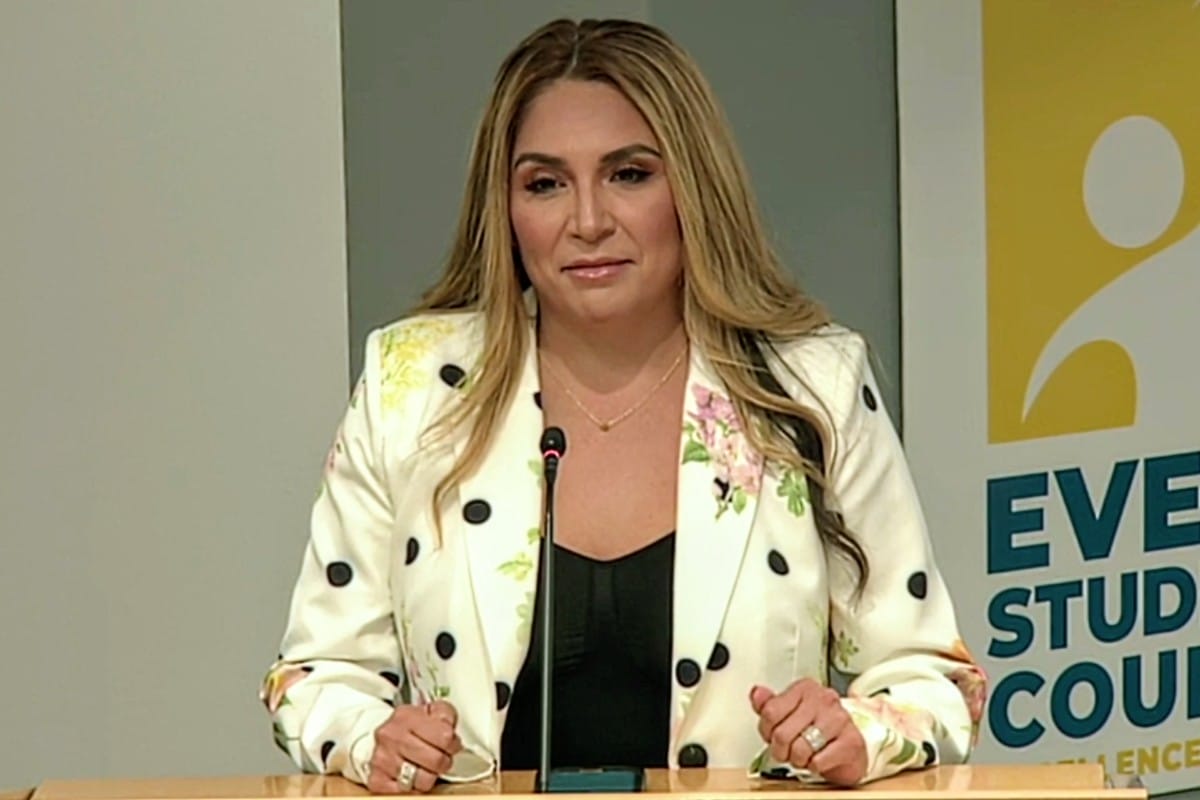 Heidi Vega speaks to School Board members (screenshot via APS) | ARLnow Woman standing behind a wooden lectern with a microphone, wearing a white floral blazer and black top, presenting at a conference.