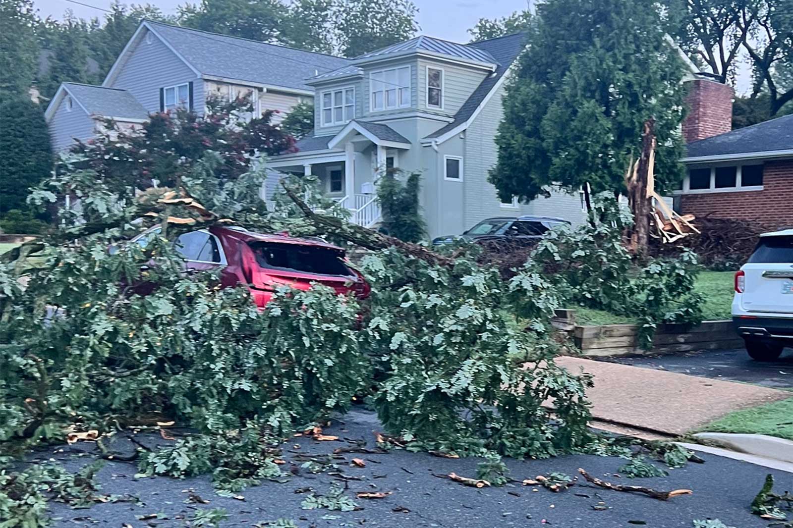 Large tree branches and debris cover a red SUV parked on a suburban street after a storm, with houses and another car in the background.