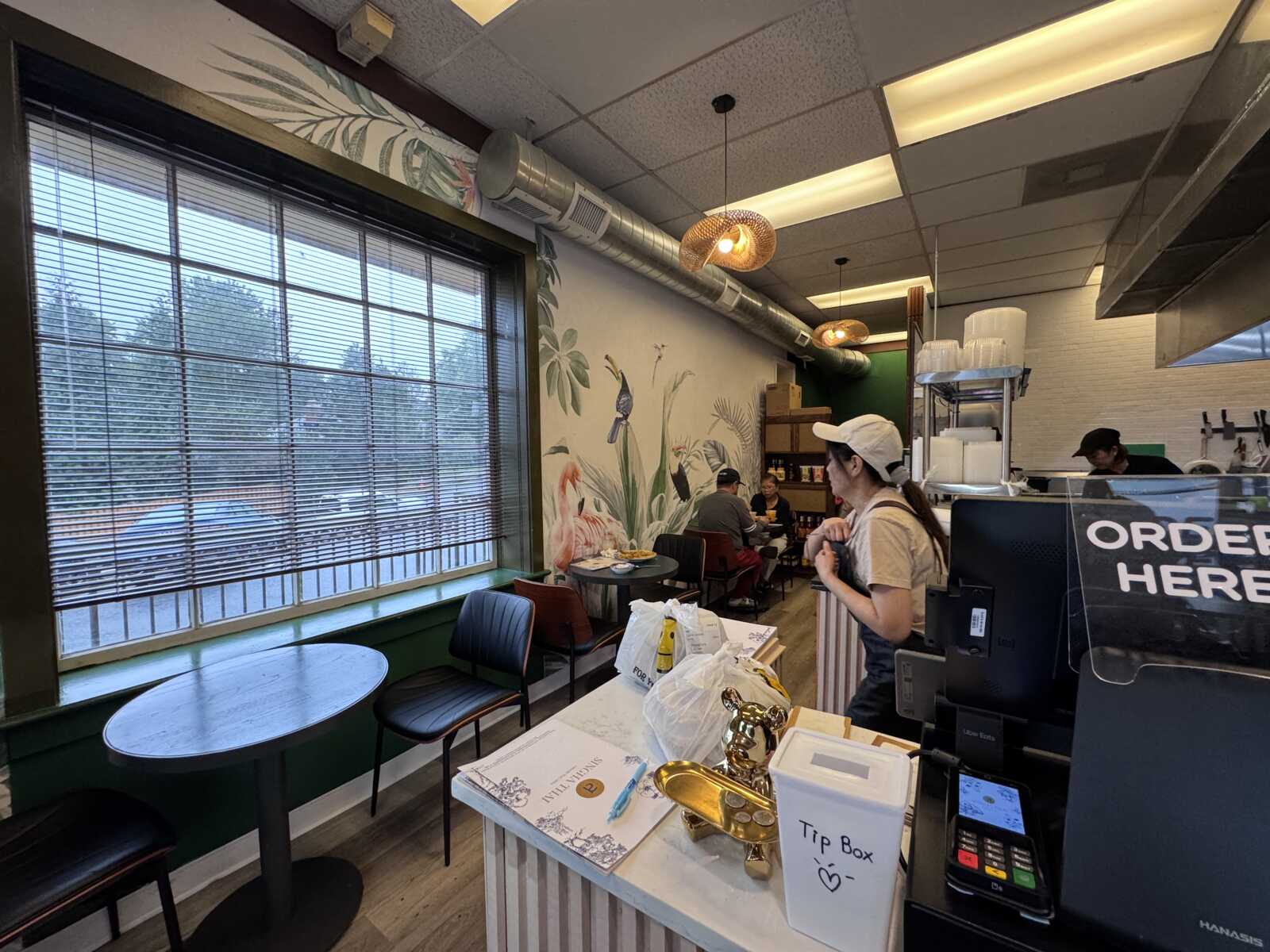 Cafe interior with a tropical plant mural along the wall, large window with blinds, and round tables with chairs; customers sit and eat while a worker stands at the counter nearby and a tip box is on the counter edge.