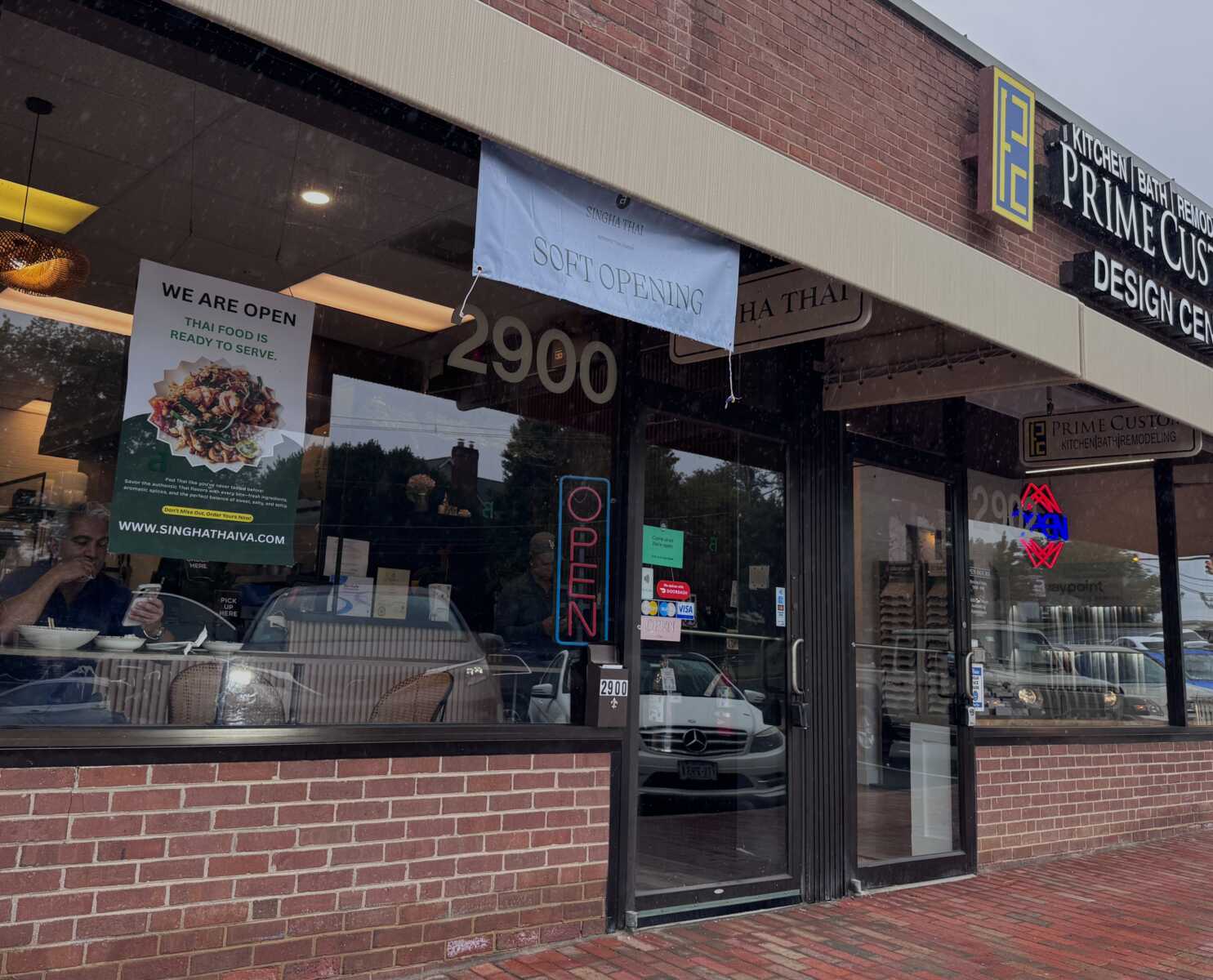 Restaurant storefront with a 'Soft Opening' banner, large glass windows, and a bright 'OPEN' neon sign; diners visible inside.