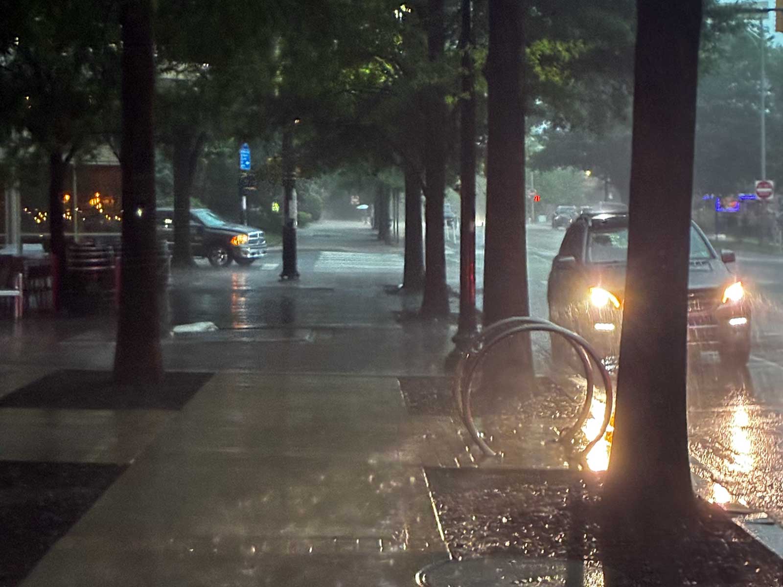 A soaking rain in Courthouse (courtesy George Brazier) | ARLnow Rainy urban street at night with car headlights and reflections on wet pavement, trees lining the sidewalk, and a circular bike rack in the foreground