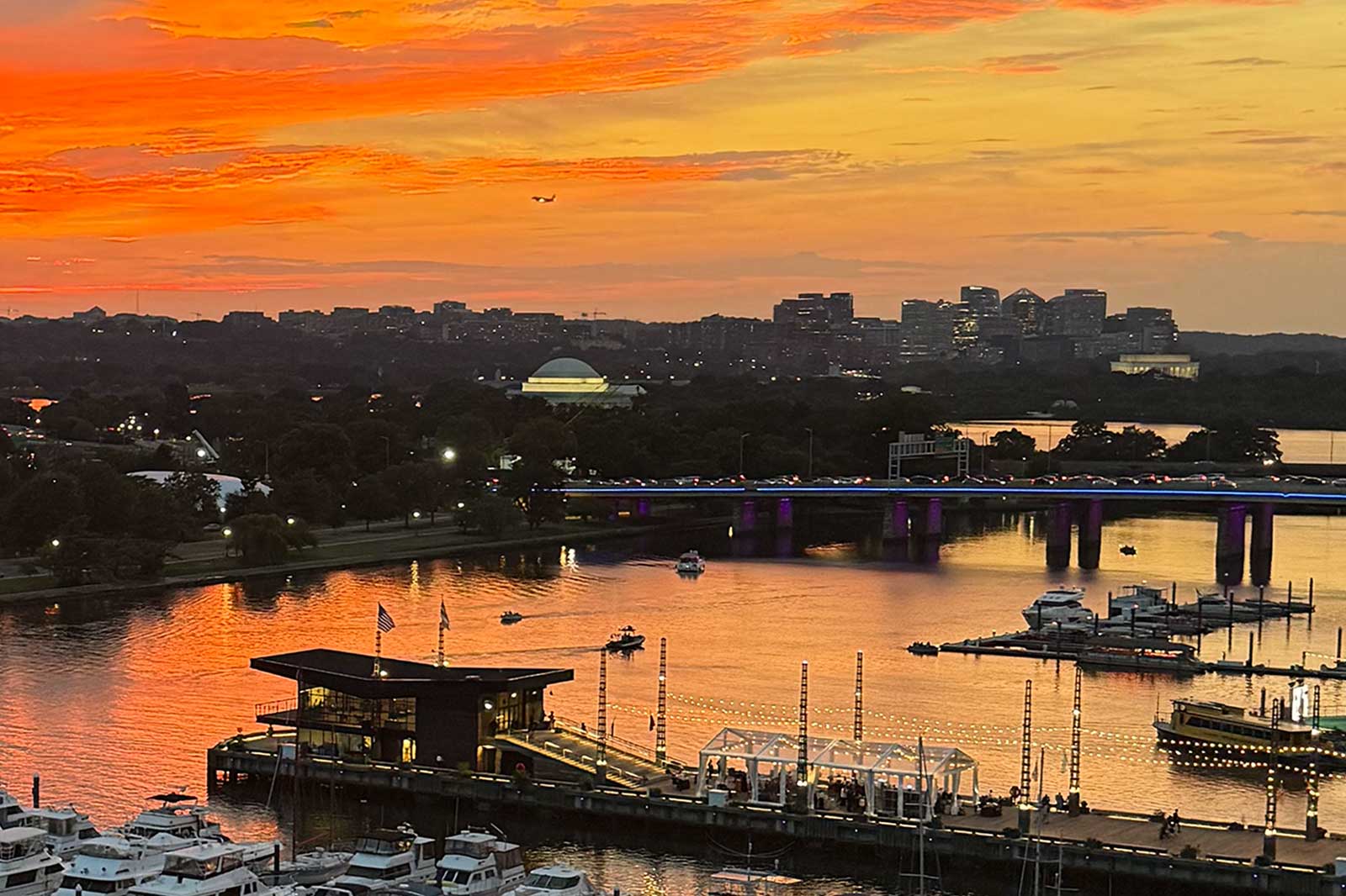 Sunset over Arlington and the Potomac, as seen from the Wharf in D.C. (courtesy photo) | ARLnow Sunset over a riverfront city with orange sky, a lit bridge, and a marina of boats along the water.