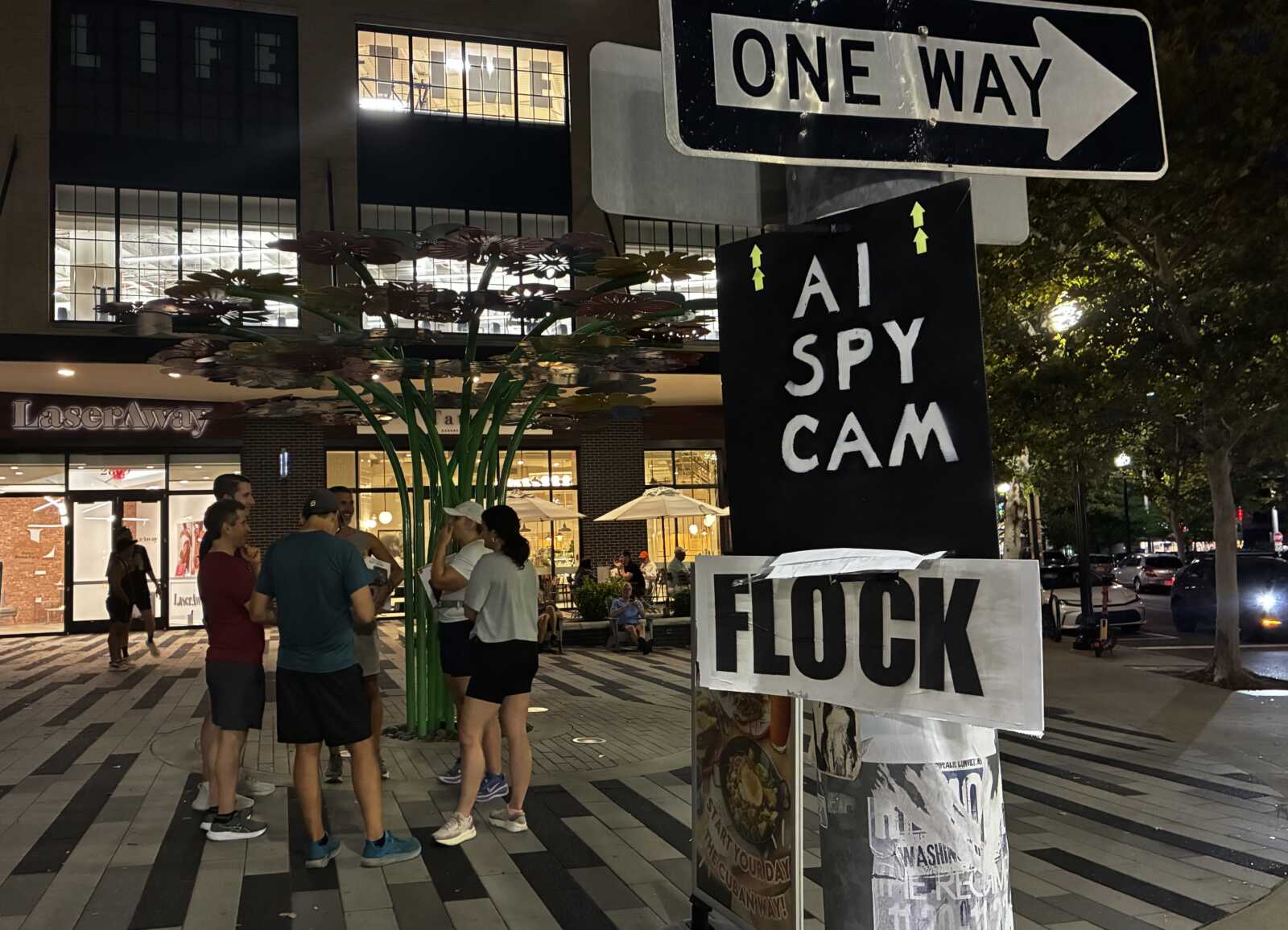 Anti-Flock sign in Clarendon (courtesy George Brazier) | ARLnow Nighttime street plaza with a group chatting near a large black banner reading'AI SPY CAM' and a white 'FLOCK' sign, plus a prominent 'ONE WAY' sign overhead.
