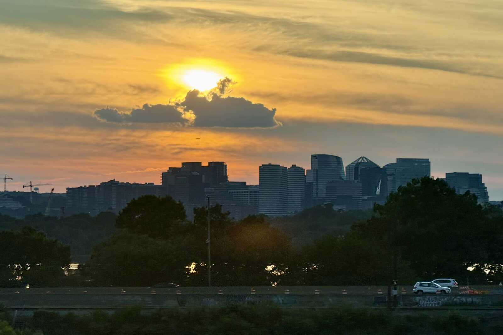 Clouds and sun over Rosslyn and the 14th Street Bridge (staff photo) | ARLnow Sunset over a city skyline with orange sky, silhouetted buildings, and trees in the foreground; a highway runs along the bottom edge.