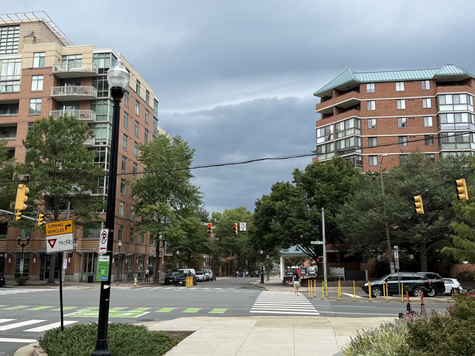 A storm front approaches in Ballston (staff photo) | ARLnow Urban intersection with brick apartment buildings, trees, and a crosswalk under a cloudy sky.