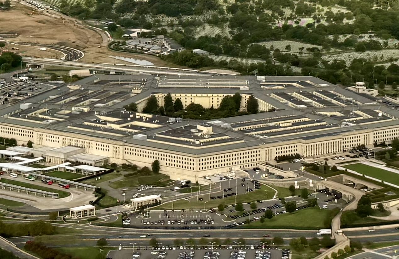 Aerial view of the Pentagon (staff photo) | ARLnow Aerial view of a large, beige, U-shaped government building with a central courtyard and extensive surrounding parking lots and roads.