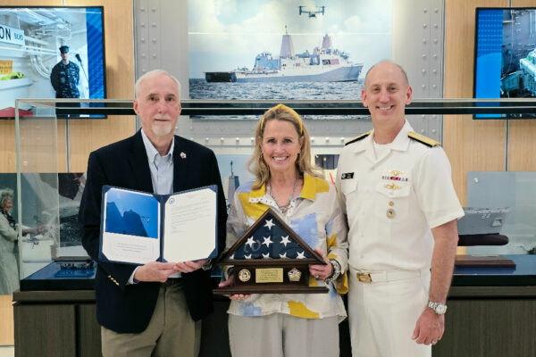 Three adults pose for a photo at an awards ceremony; the woman in the center holds a folded flag display while the man on the left shows a certificate, and a smiling sailor stands on the right with medals visible.