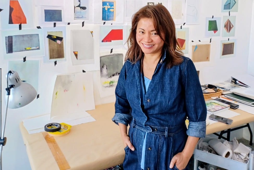 Local artist Courtney Nguyen in her studio (photo courtesy Melanie Kehoss) | ARLnow Woman in a denim jumpsuit standing in a bright art studio, wall covered with taped sketches and paintings, smiling.