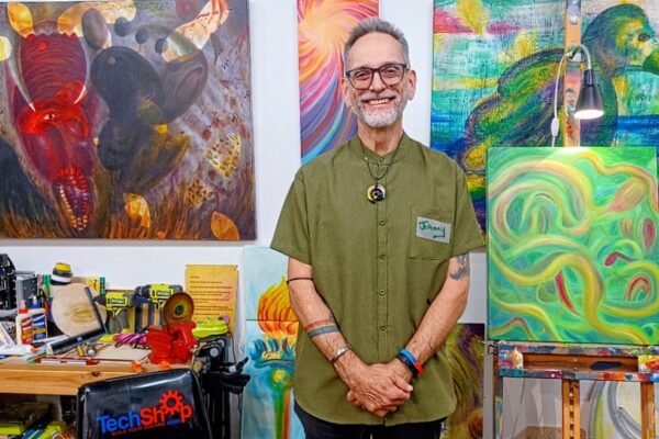 Artist Johnny stands in his studio, smiling, with colorful abstract paintings and painting tools in the background.