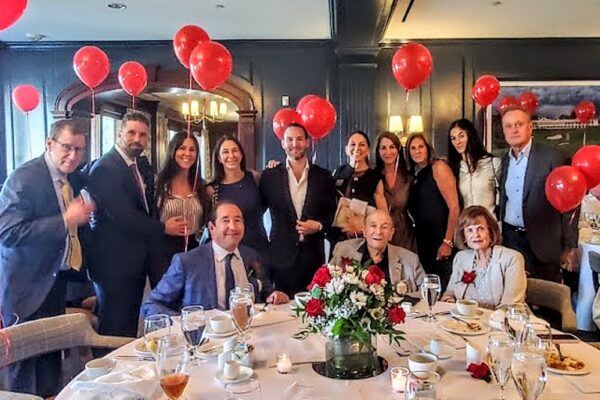 Group of adults in formal wear posing around a round banquet table with a red-and-white floral centerpiece and red balloons, smiling for a photo.