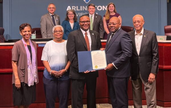 Eight adults in formal attire pose in a council chamber; center man receives a certificate from a colleague while others smile behind.