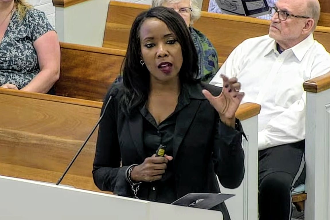 Northern Virginia Transportation Authority CEO Monica Backmon (screenshot via City of Falls Church) | ARLnow Black woman in a black blazer speaks at a podium in a meeting room, gesturing with her right hand as she addresses an audience.