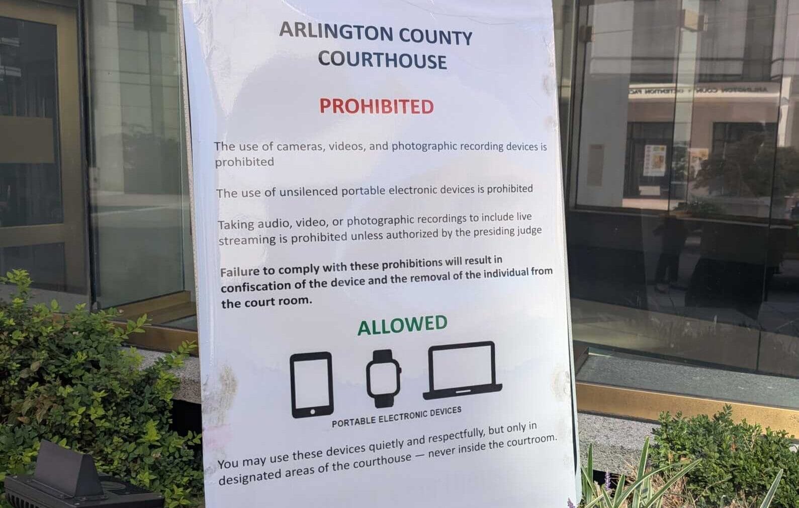 Sign outside Arlington County Courthouse listing prohibited devices (cameras, video, recording) and allowed portable electronic devices with icons on the bottom.