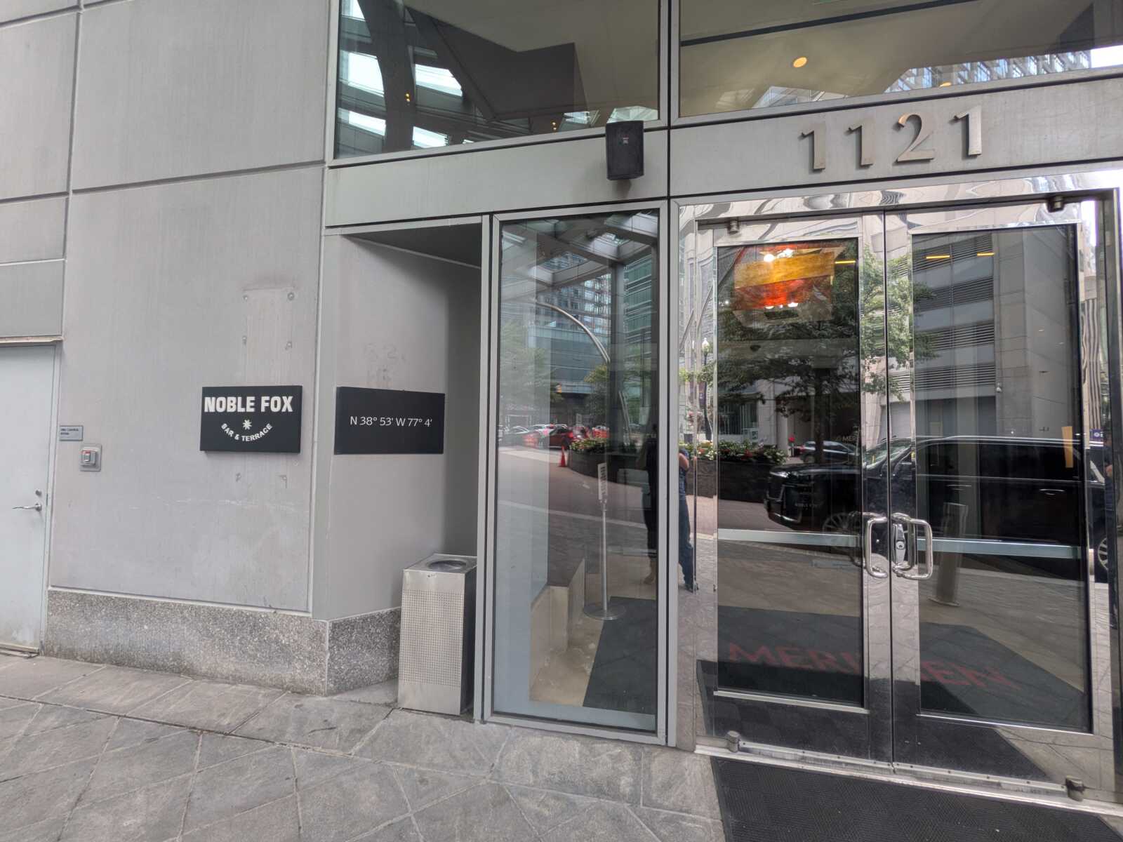 Entrance to a modern building with glass doors and the number 1121 above it, reflecting the street and parked cars outside. See signs for Noble Fox Bar & Terrace on the left wall.