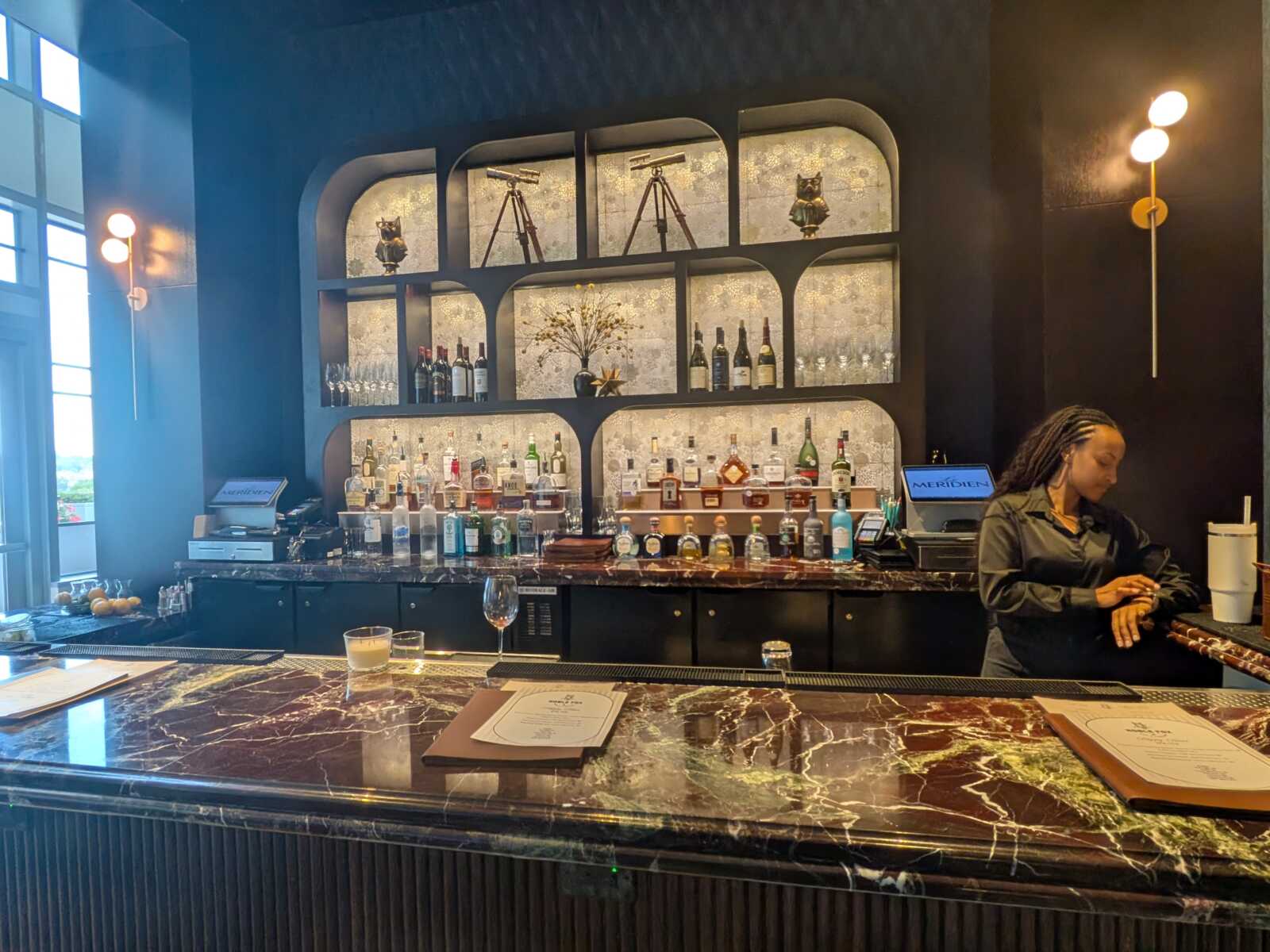 Dark bar interior with a marble counter and arched, backlit shelves holding bottles and glassware; a bartender stands at the right preparing a drink.