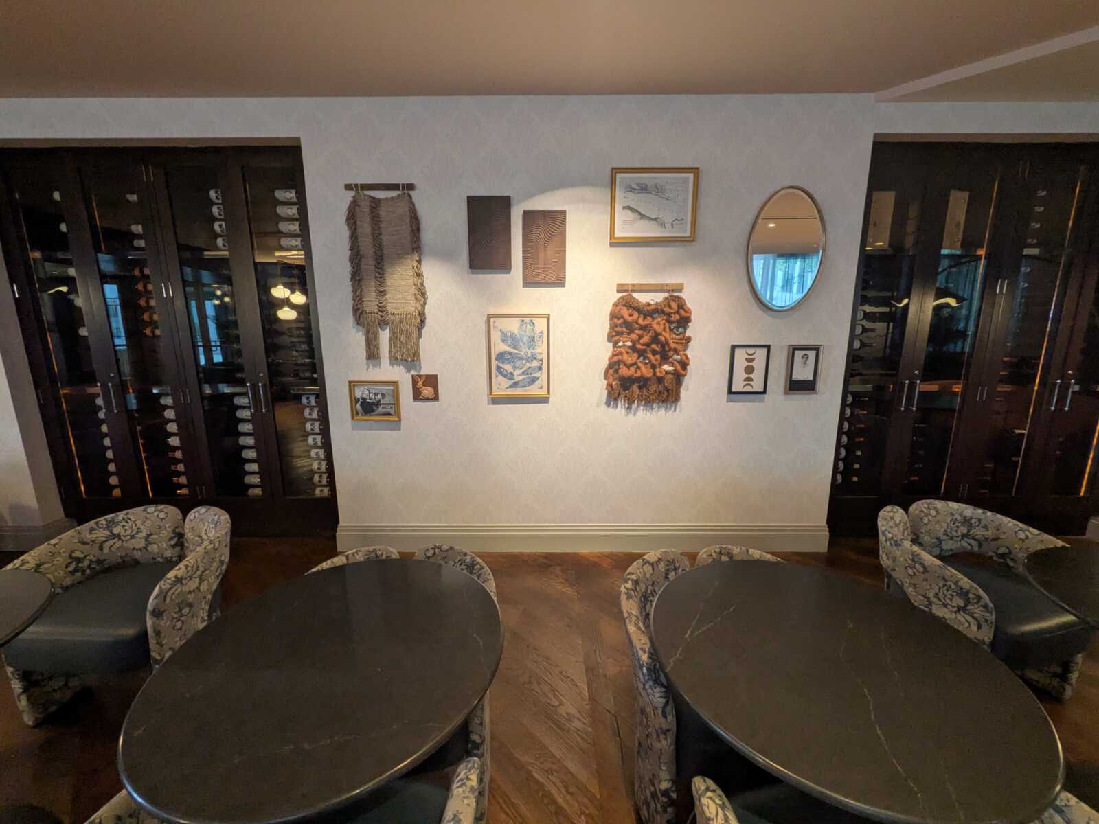 Cozy wine lounge with dark wood wine cabinets lining both sides, round black tables, and floral upholstered chairs; wall displays include woven textile pieces, framed art, and an oval mirror.