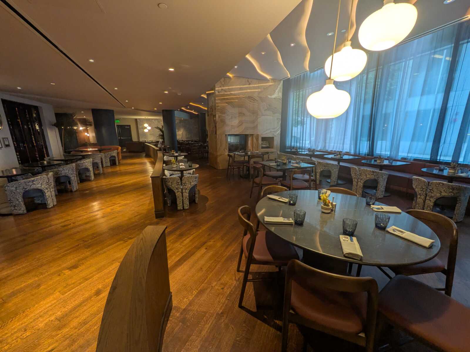 Upscale restaurant dining area with round tables, patterned chairs, and large pendant lights by a blue window wall.